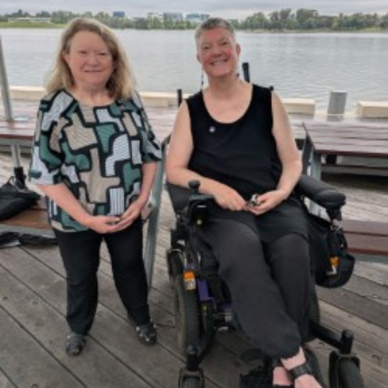 Two women smiling together on a timber waterfront deck, one standing and one using a power wheelchair, with a river and city skyline behind them